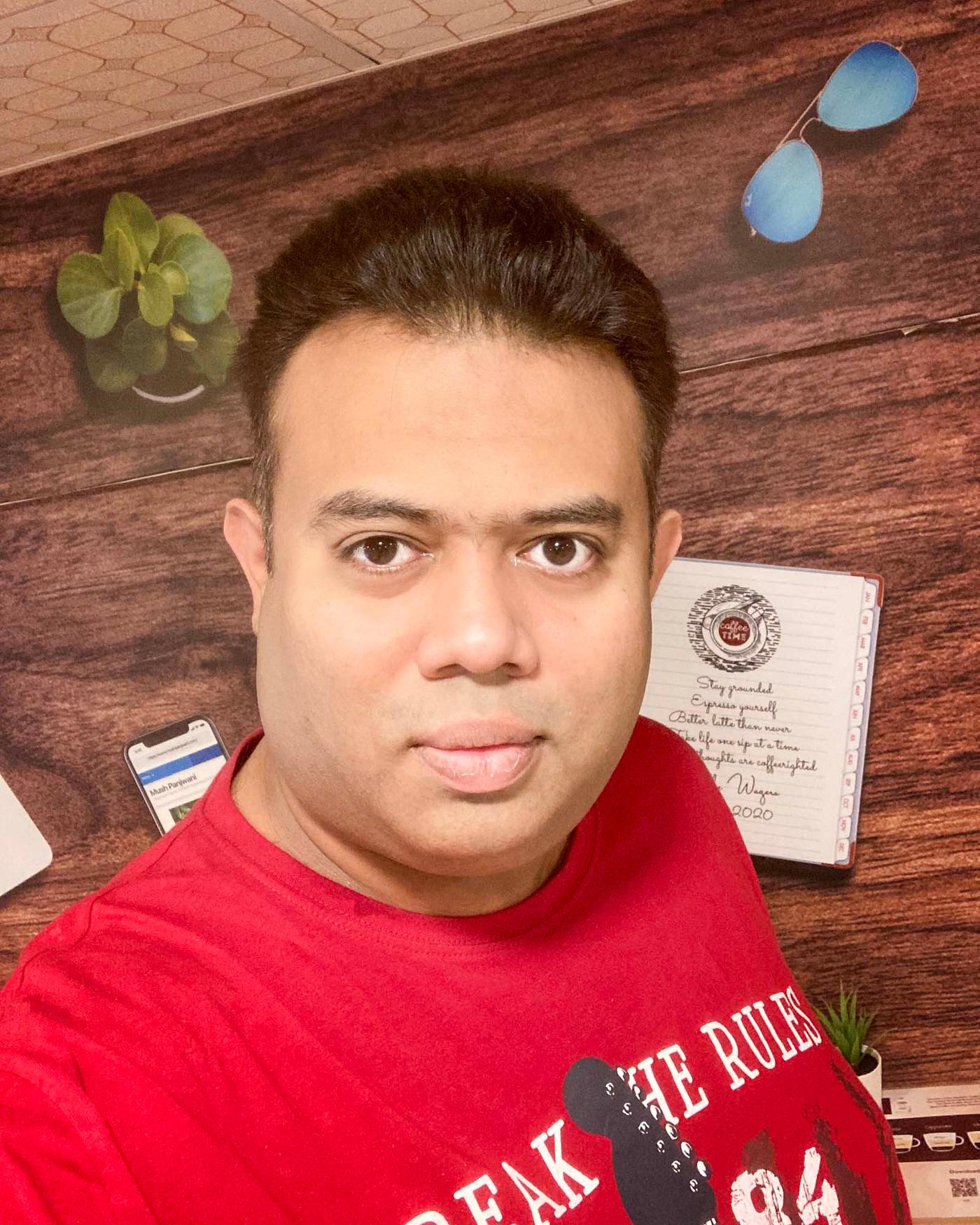 A man in a red T-shirt looks at the camera. In the background is a wood wall.