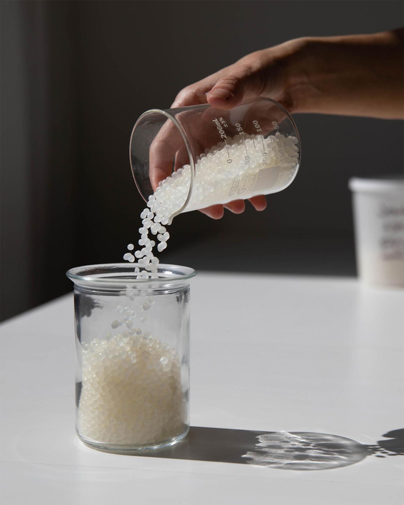 Tiny round white beads being poured from one glass vessel into another on a white laboratory-like surface.