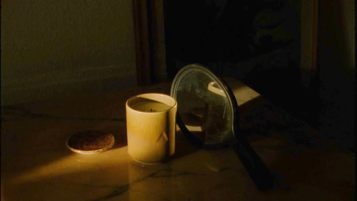 A candle in a ceramic container sits on a marble table.  A mirror reflects the light.