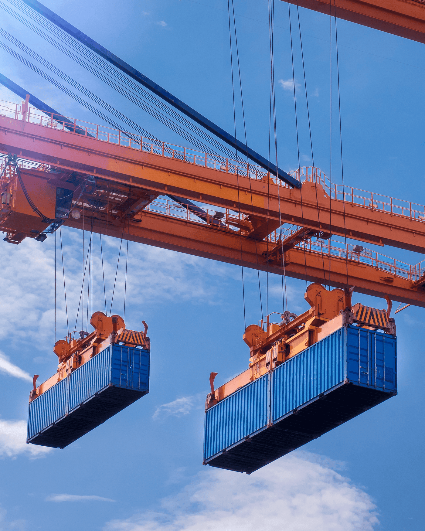 Two blue shipping containers shown from below, being lifted in the air by a large red crane.