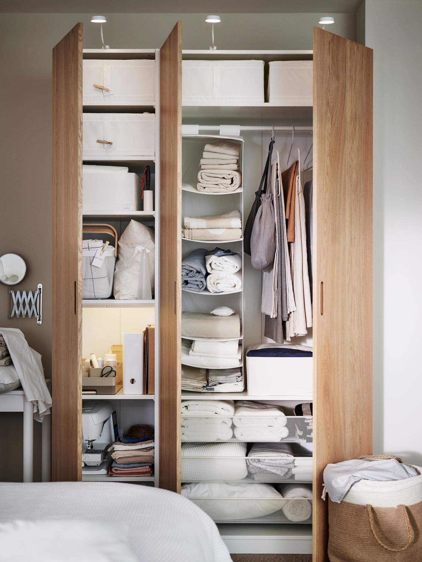 A PAX wardrobe in a bedroom, with open doors. Inside the wardrobe are shelves filled with clothes and white storage boxes and on the right hand side hang clothes.