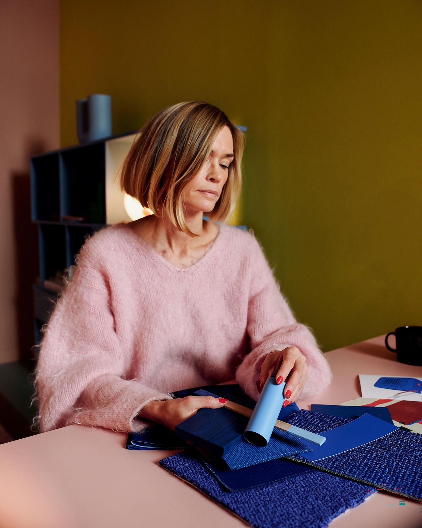 Tekla Severin sits at a desk in a pink outfit, holding a small blue-tipped stick and comparing color samples. Blue textiles and sketches are visible on the desk.