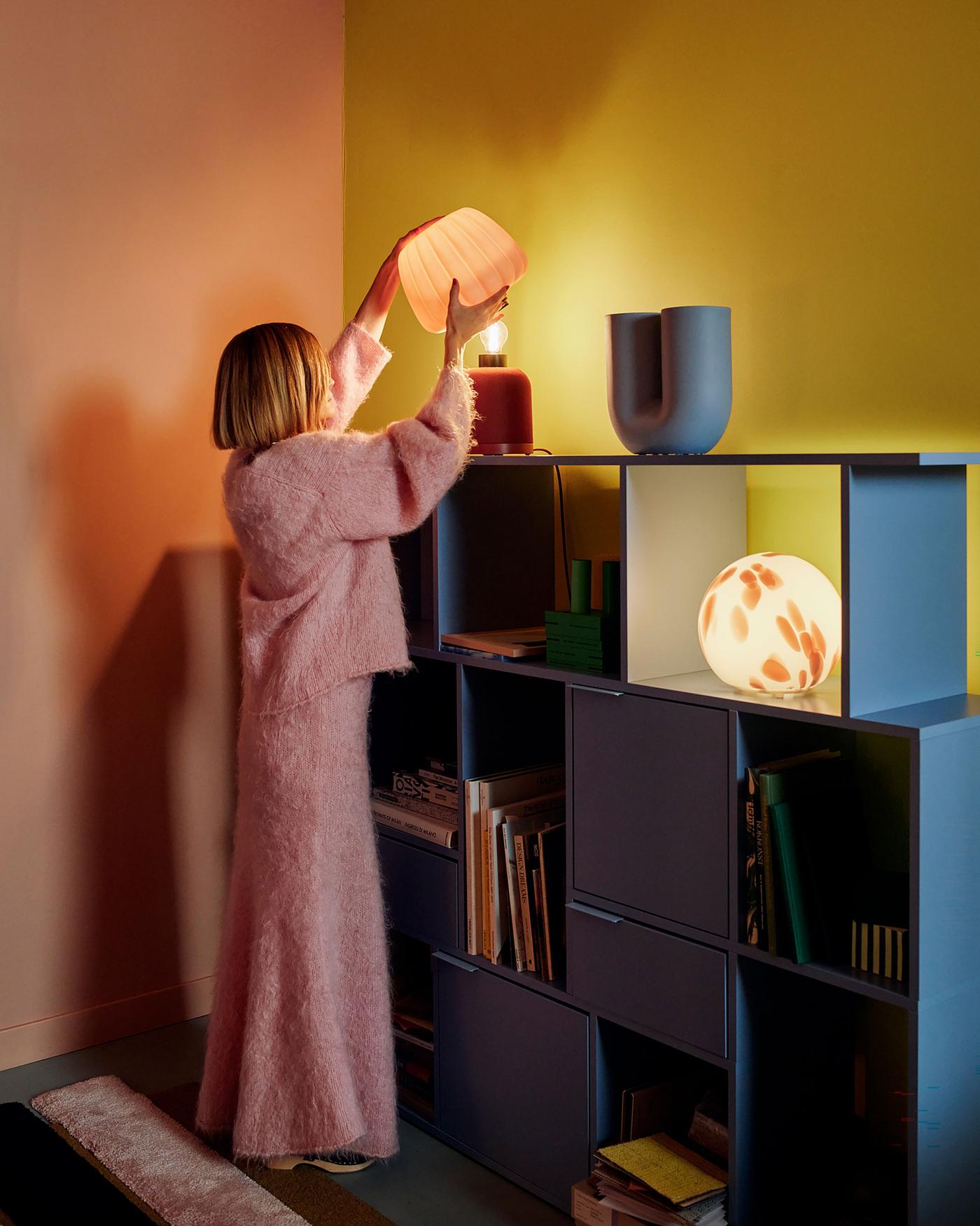 Tekla Severin stands in her studio, facing a blue bookshelf with yellow walls in the background. She lifts a pink lampshade from a red KULGLASS speaker. A spherical FADO lamp is visible on the shelf below. 