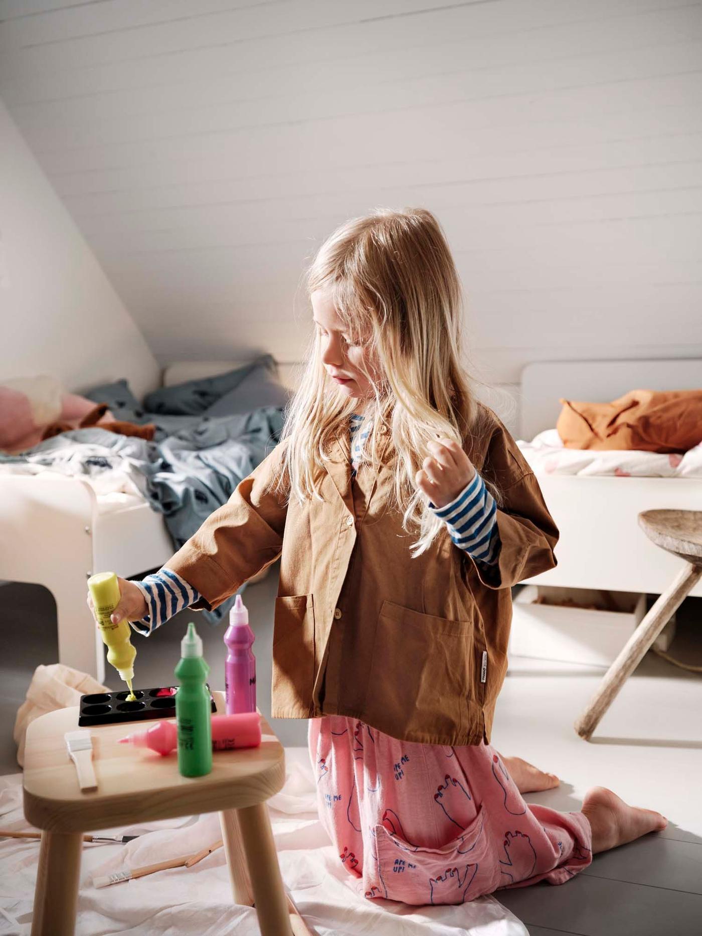 A girl kneels on the floor of her bedroom. She is squeezing a bottle of paint onto a palette. She's holding her hair.