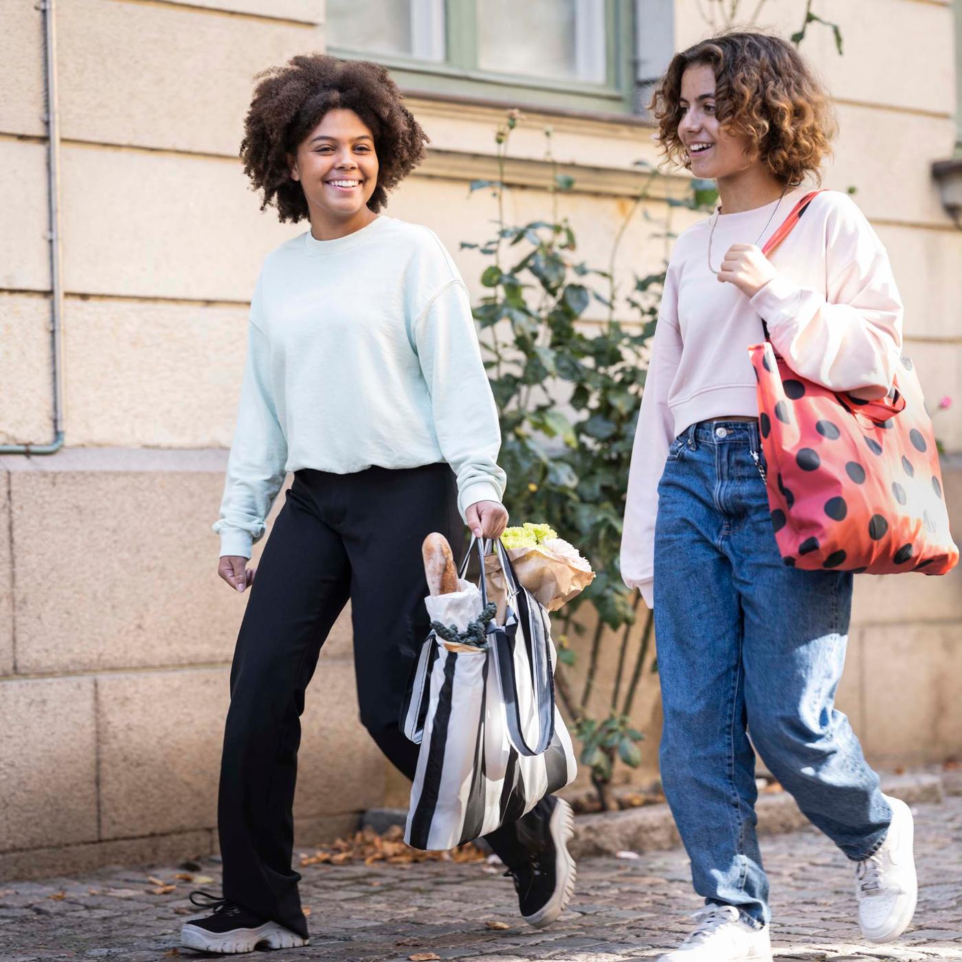 Two people walk along a sidewalk. The person on the left of the image is carrying a striped/black white SKYNKE carrier bag.