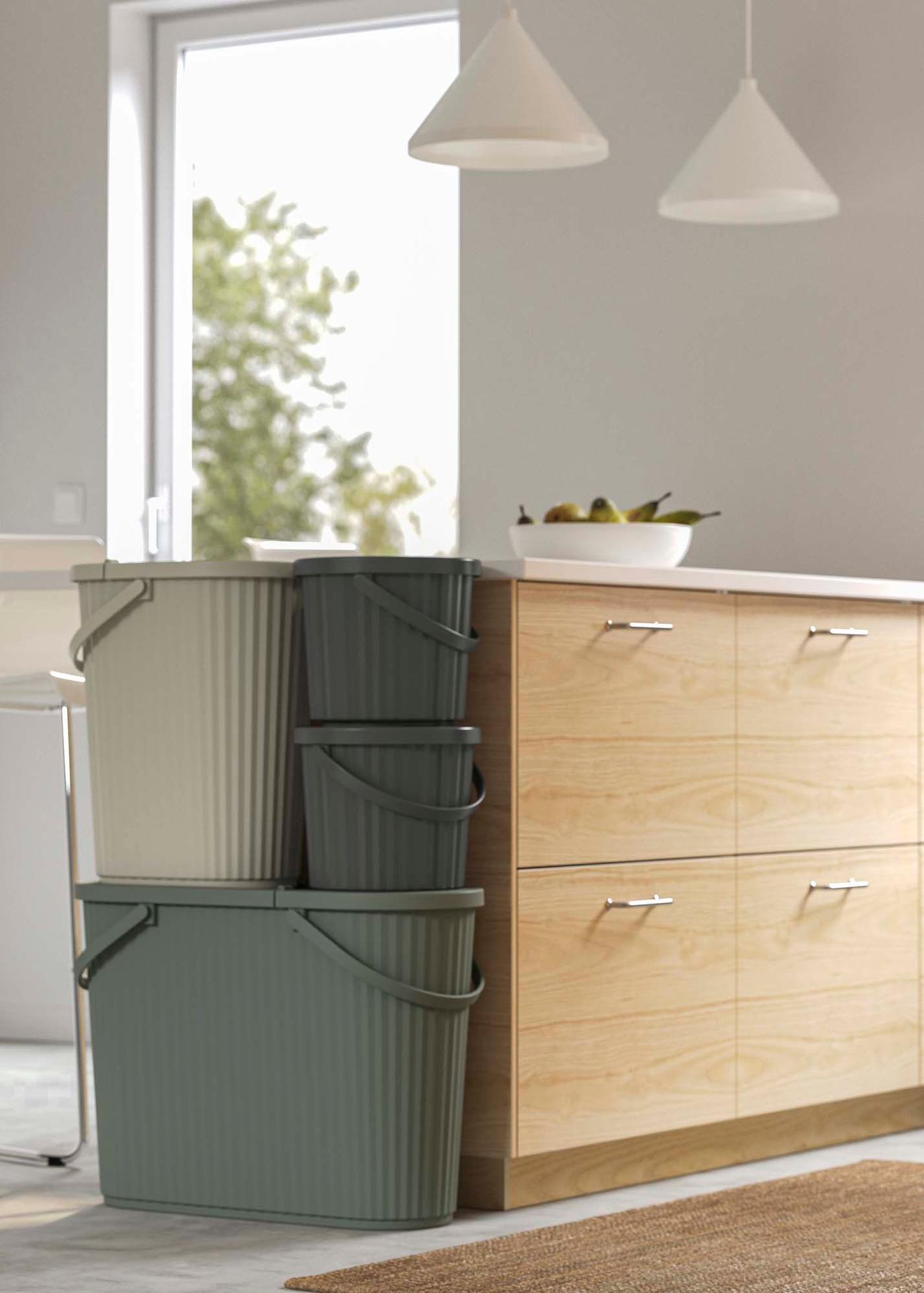 Three recycling bins in different shades of green are stacked against a wood kitchen island in a white kitchen.