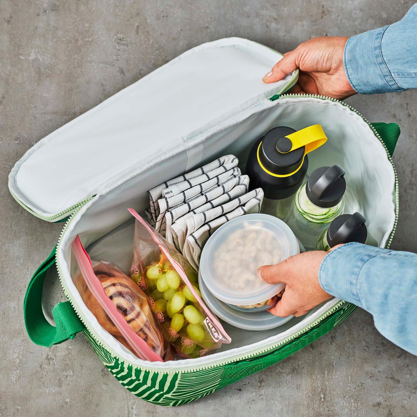 A pair of hands reaching into a patterned/green NÄBBFISK cooler bag, filled with picnic items.