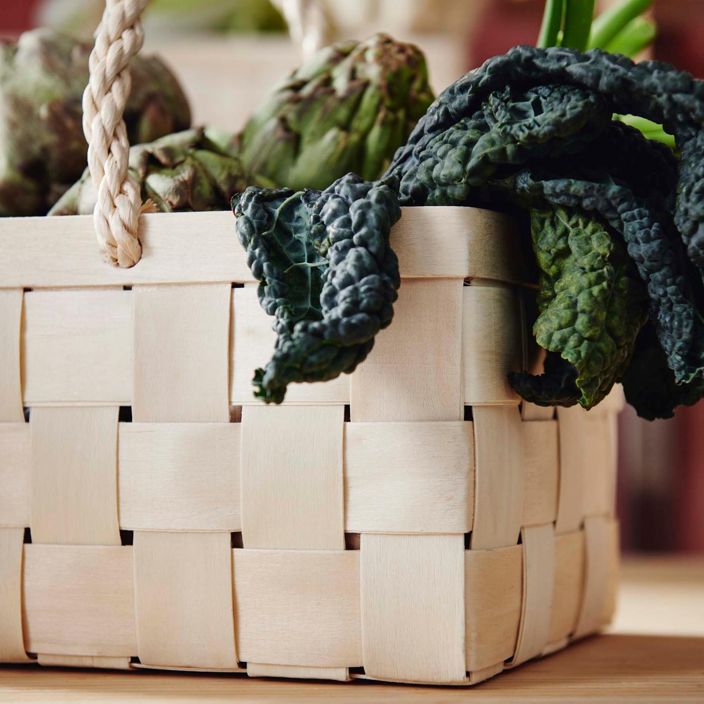 A close-up cropped image of a woven wood KÖSSEBÄR basket filled with kale and artichokes.