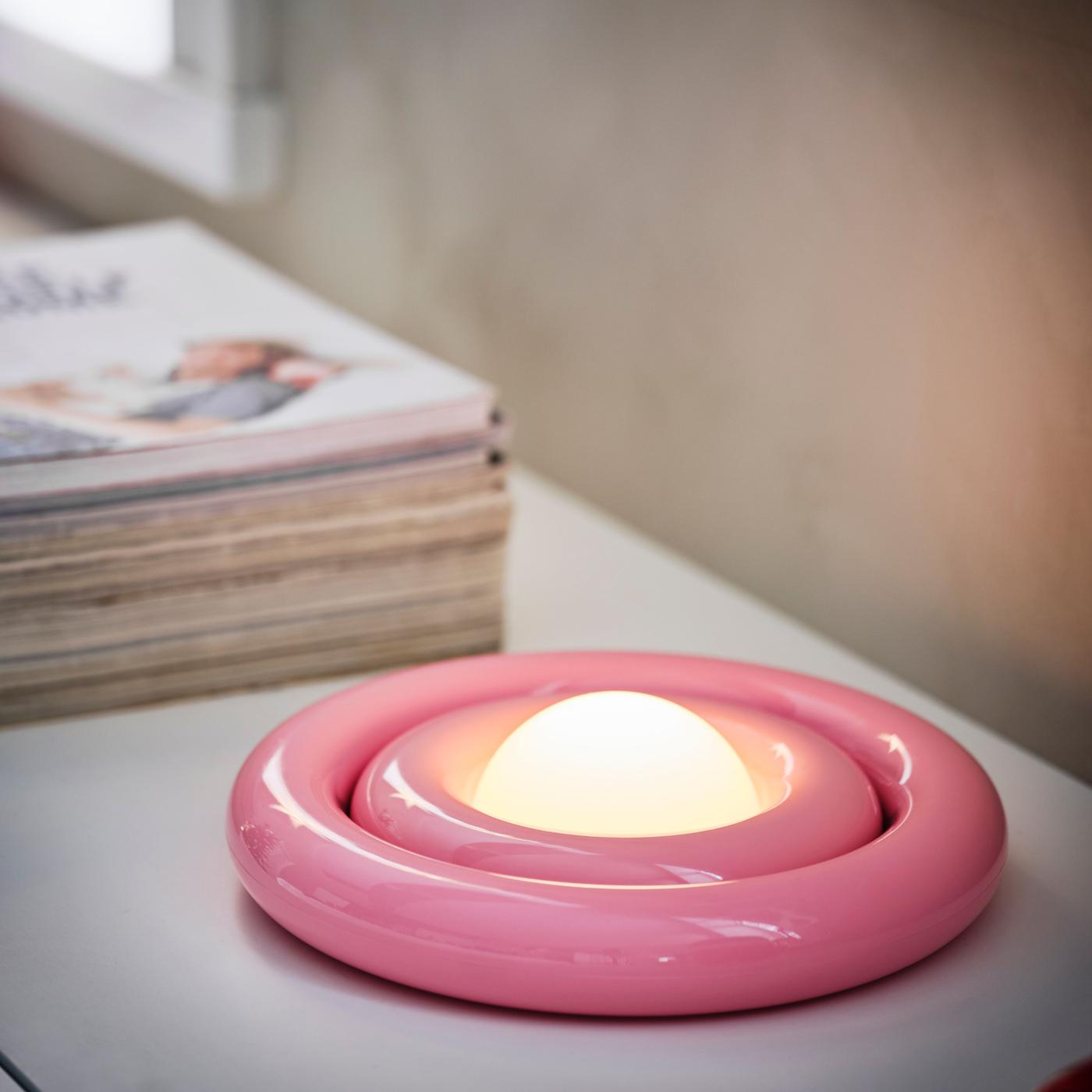 Pink round lamp with layered, swirl-like design, lying flat on a surface, lit and portable.