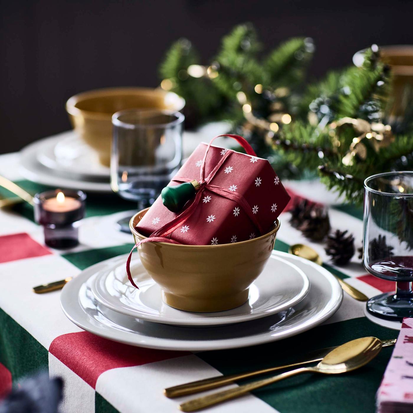 A close-up of a Christmas table setting featuring a bold patterned tablecloth and a small wrapped gift placed inside a bowl.