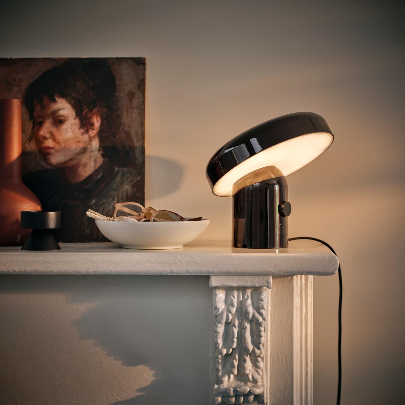 A black speaker lamp illuminated, placed on a ledge with bowls and artwork nearby against white walls.