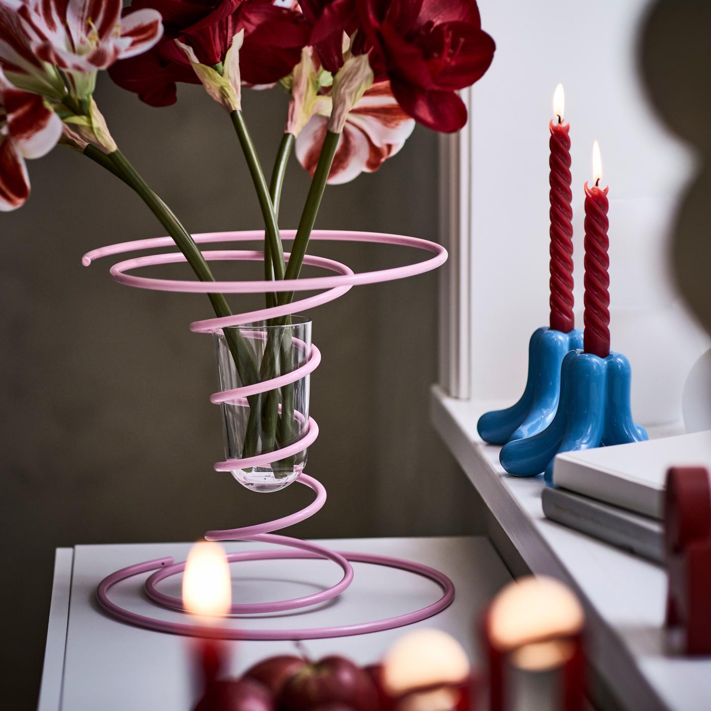 A lPink steel vase with red flowers on the windowsill, with lit red candles in blue candlesticks in the background.
