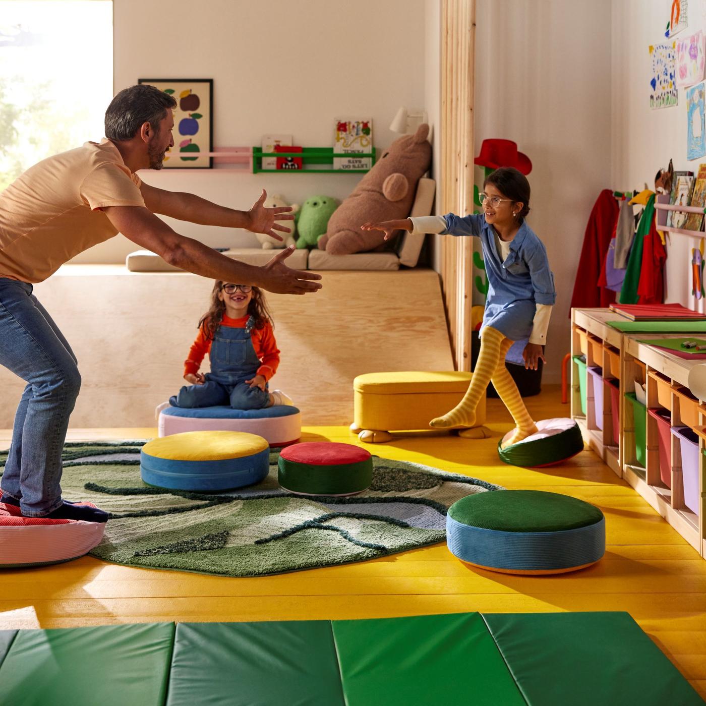 A parent and two children playing together on floor cushions and a light green rug in a bright indoor setting.
