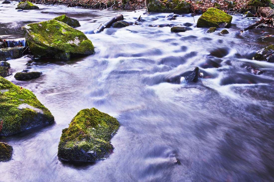A brook or shallow river. Mossy green rocks stick out of the water, as the water runs around them.