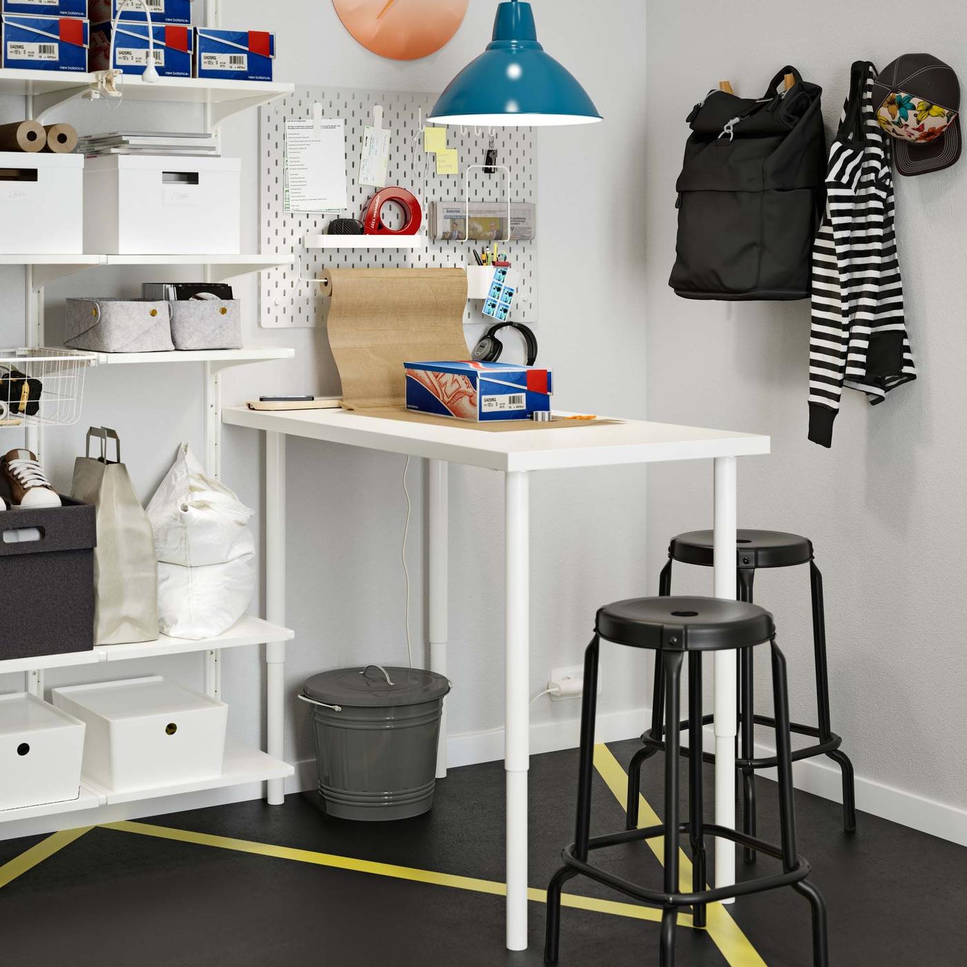 A grey bin with a lid underneath a high white table-desk in the corner of a room.