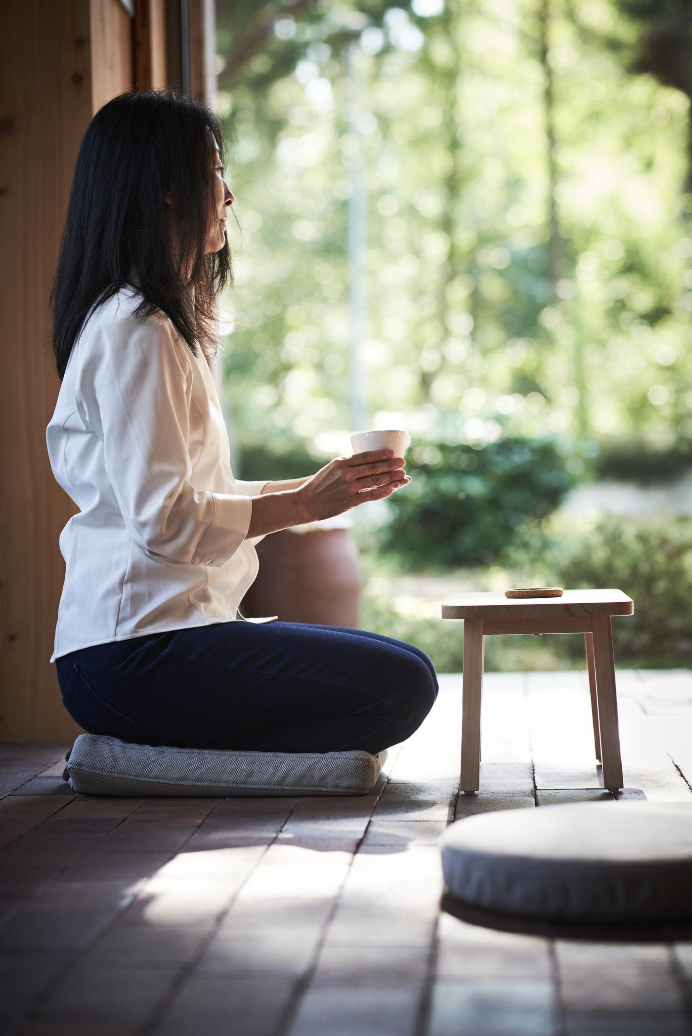 A woman holding a cup while sitting on a cushion on a terrace floor, using a stool as a table.