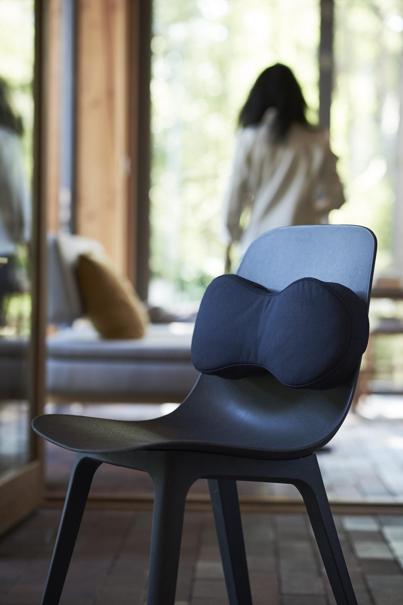 A bedroom with a chair with a back cushion. In the background there's a bed and a woman by a window.