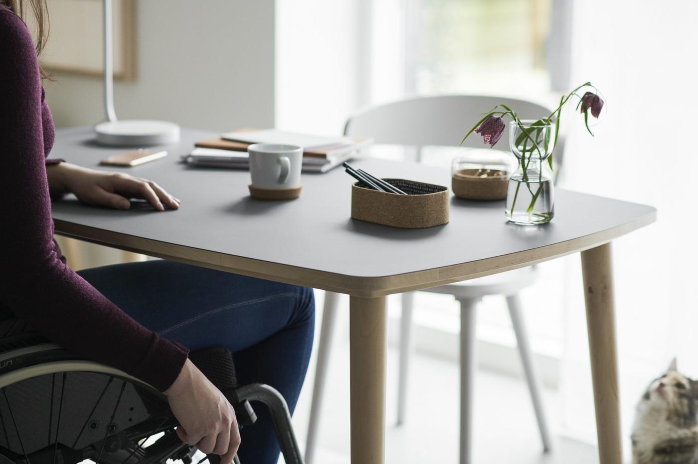 A woman in a wheelchair sitting at a table with a cup of coffee, notebooks, pens, a vase with flowers, and a cat.