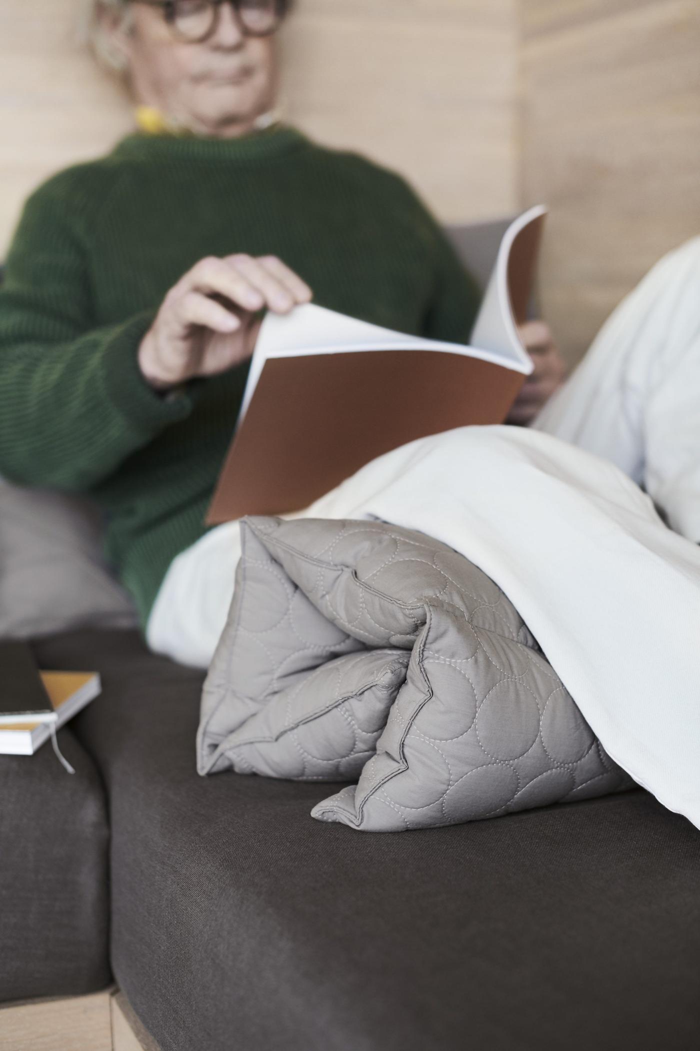 A man laying on a couch while reading a book with a duvet rolled up for support under his knees.