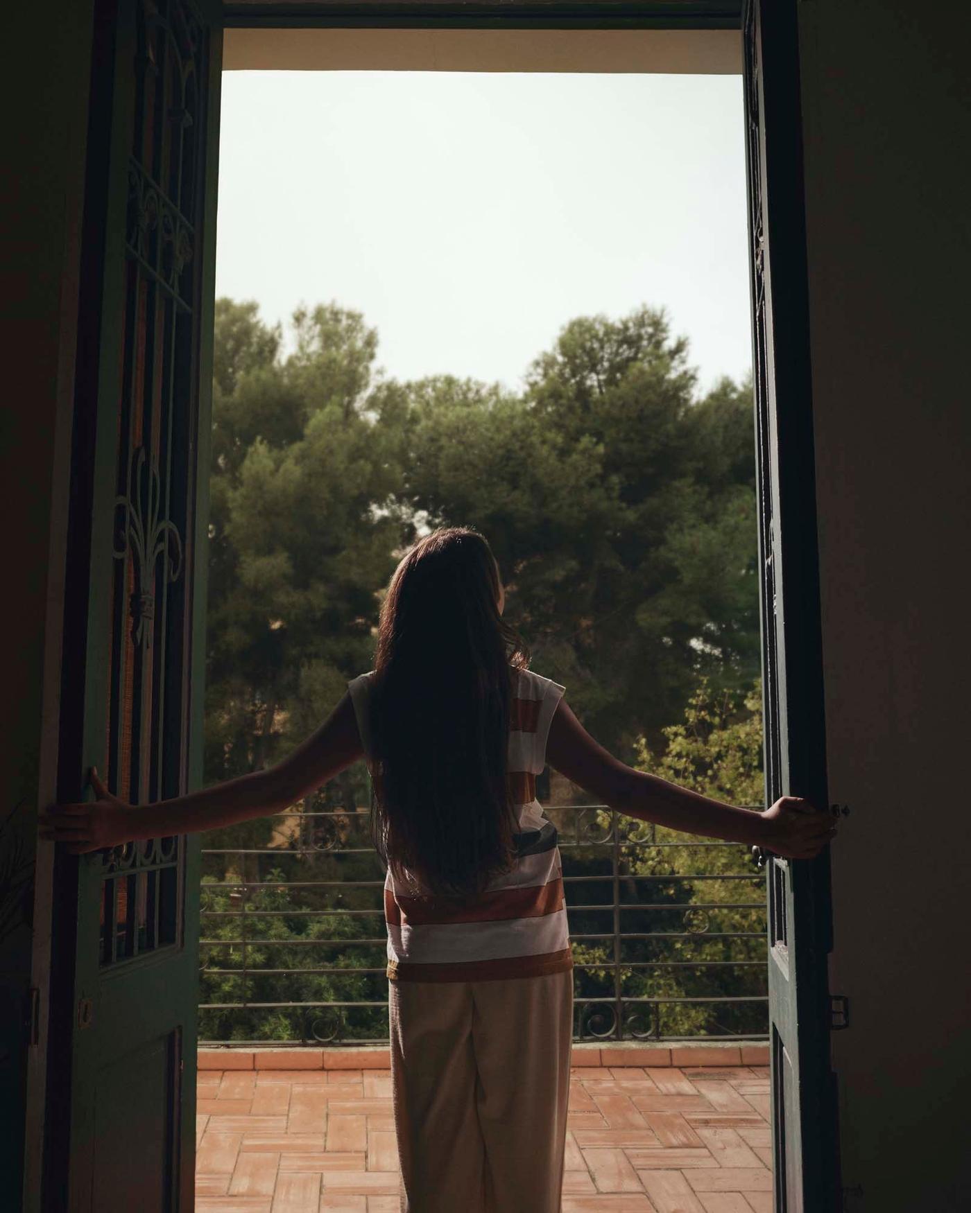 A person stands in a room, in front of an open window. Taken from behind the image shows the person's view of trees and sunny sky.