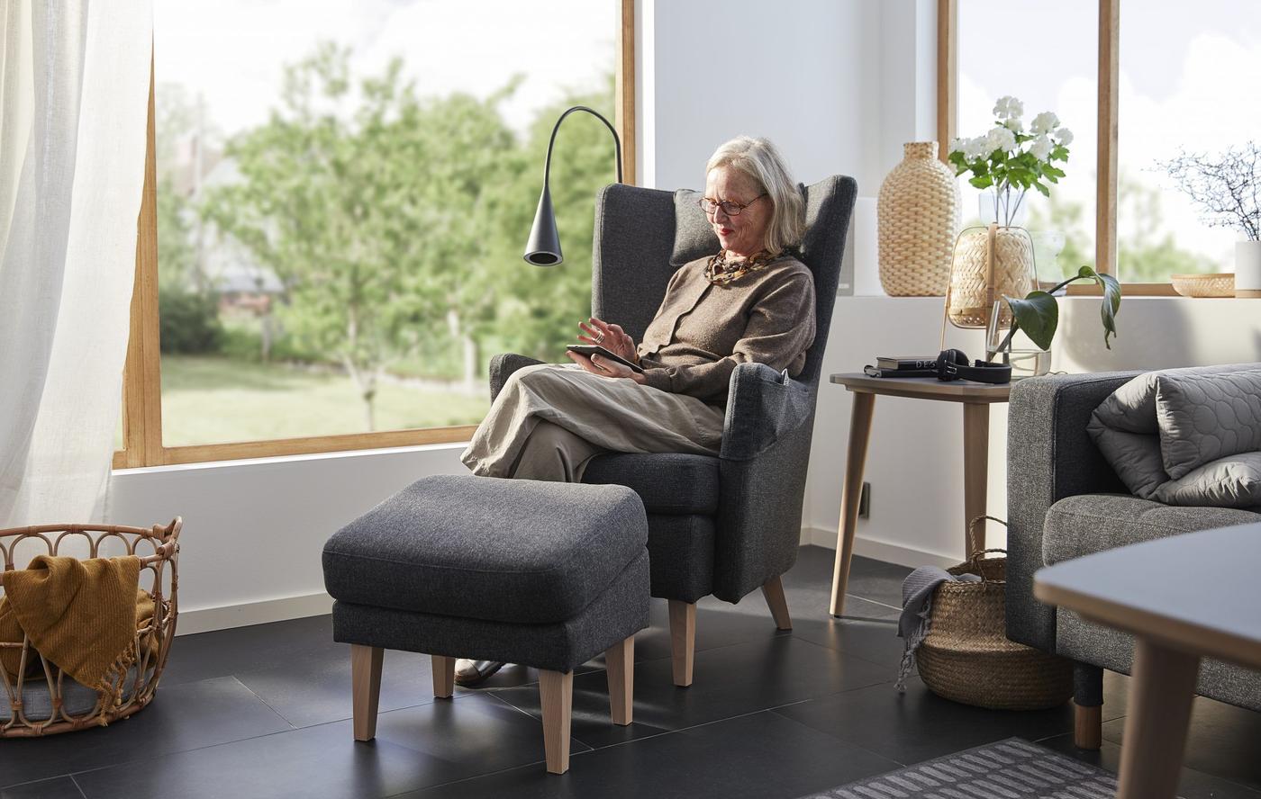 A woman sitting in a wing chair with a foot stool in a Scandi style room while using a smart device.