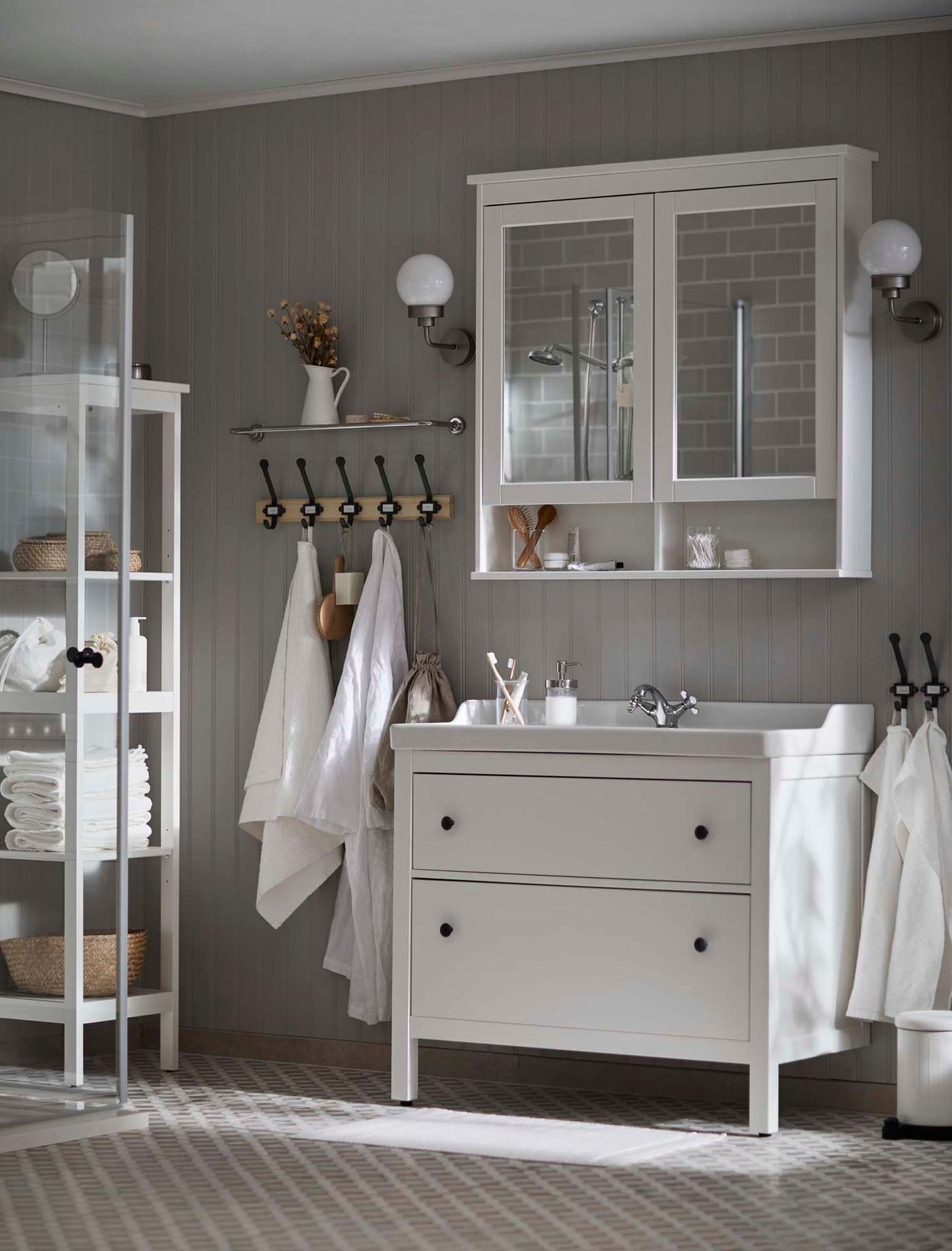 A grey bathroom with white furniture. A cabinet stands above the washbasin, with hooks and a storage unit nearby.
