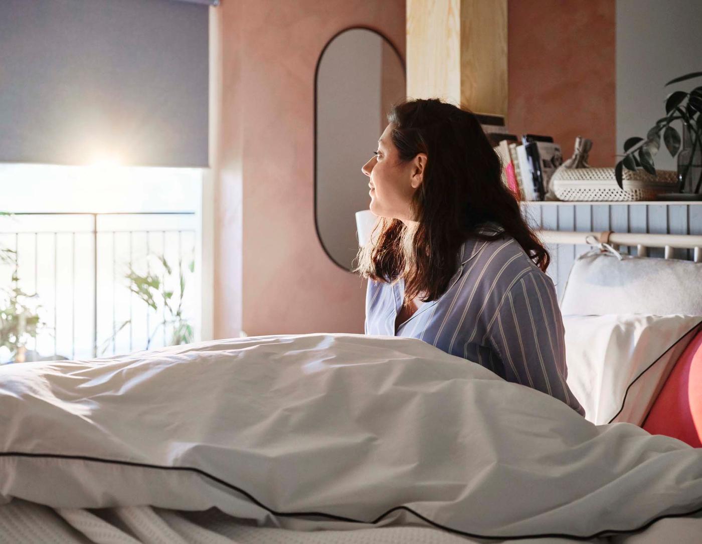 A person sits up in bed, looking out of the window as the sun shines through.