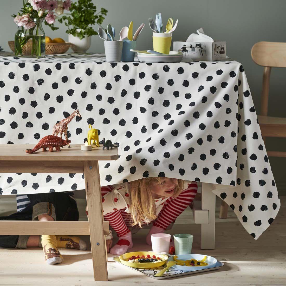 A child hides under a dining table covered in a black and white spot tablecloth. She peeps out. There are plates of food and drink next to her as she waits for her friend to find her.