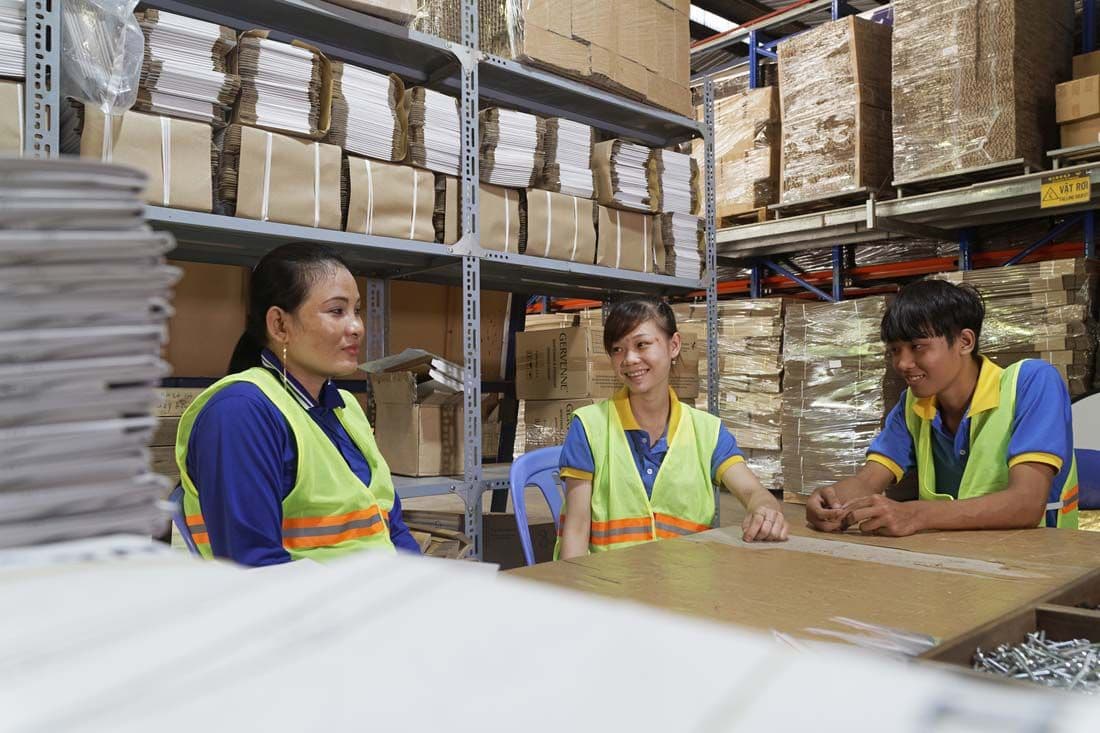 Three factory workers in conversation are sat at table with shelves of packaging behind them.