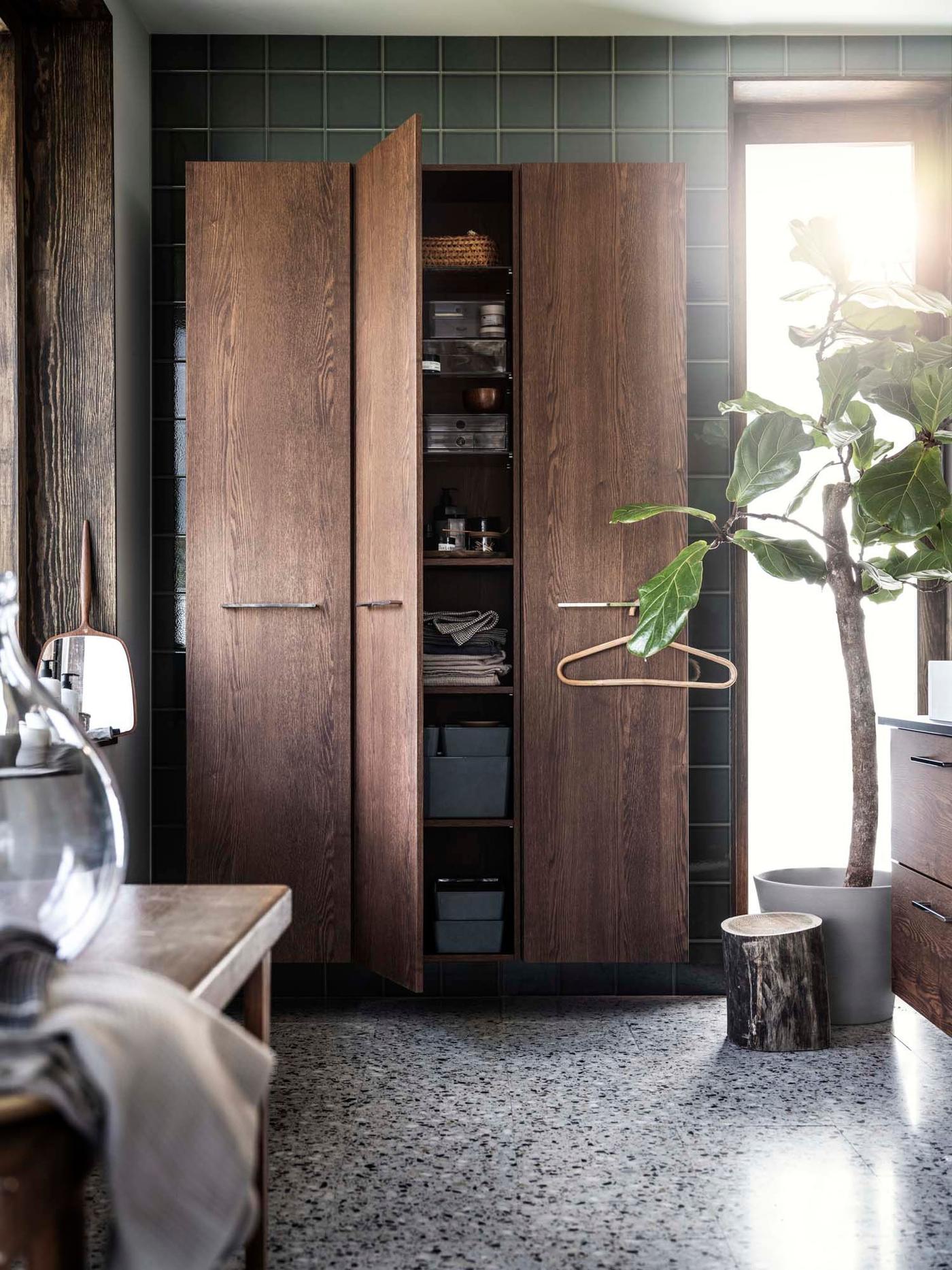 A green bathroom with a large plant and a dark wood cupboard. One door is open, revealing bathroom products.