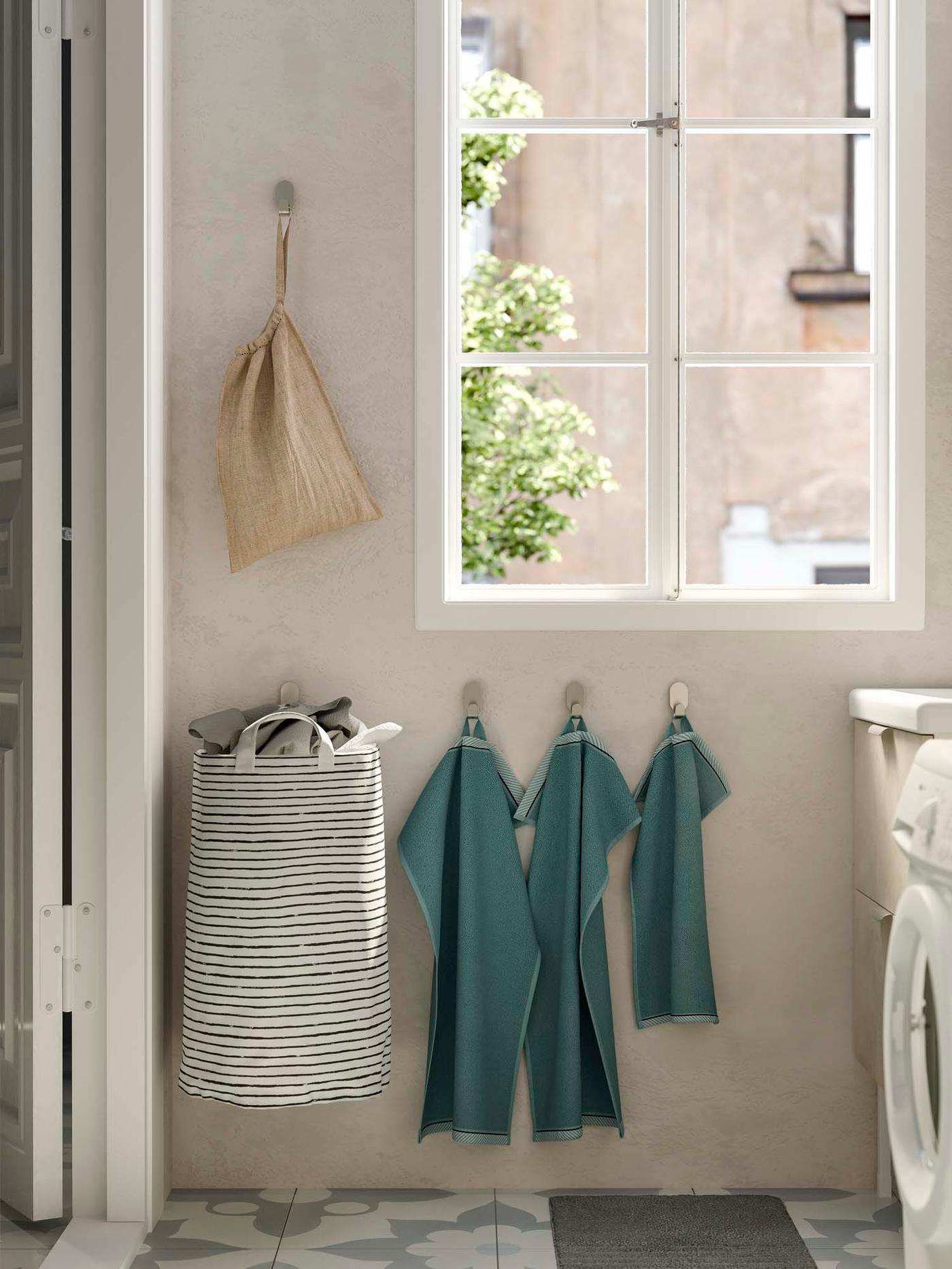 A photo of a bathroom wall with towels hanging next to a laundry bag and below a window.