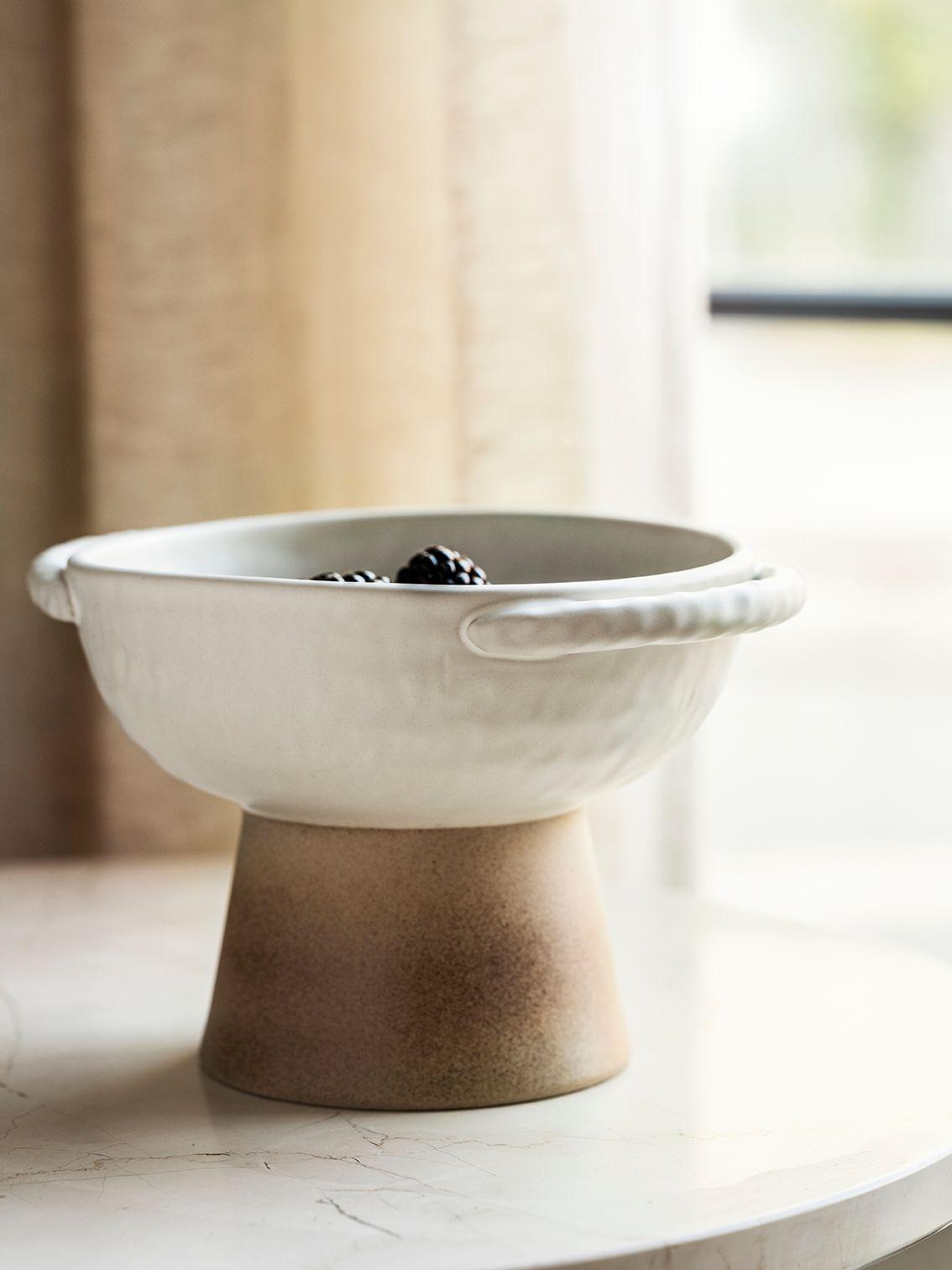 A white ceramic bowl with a brown base, on a white marble tabletop by a window with beige curtains.