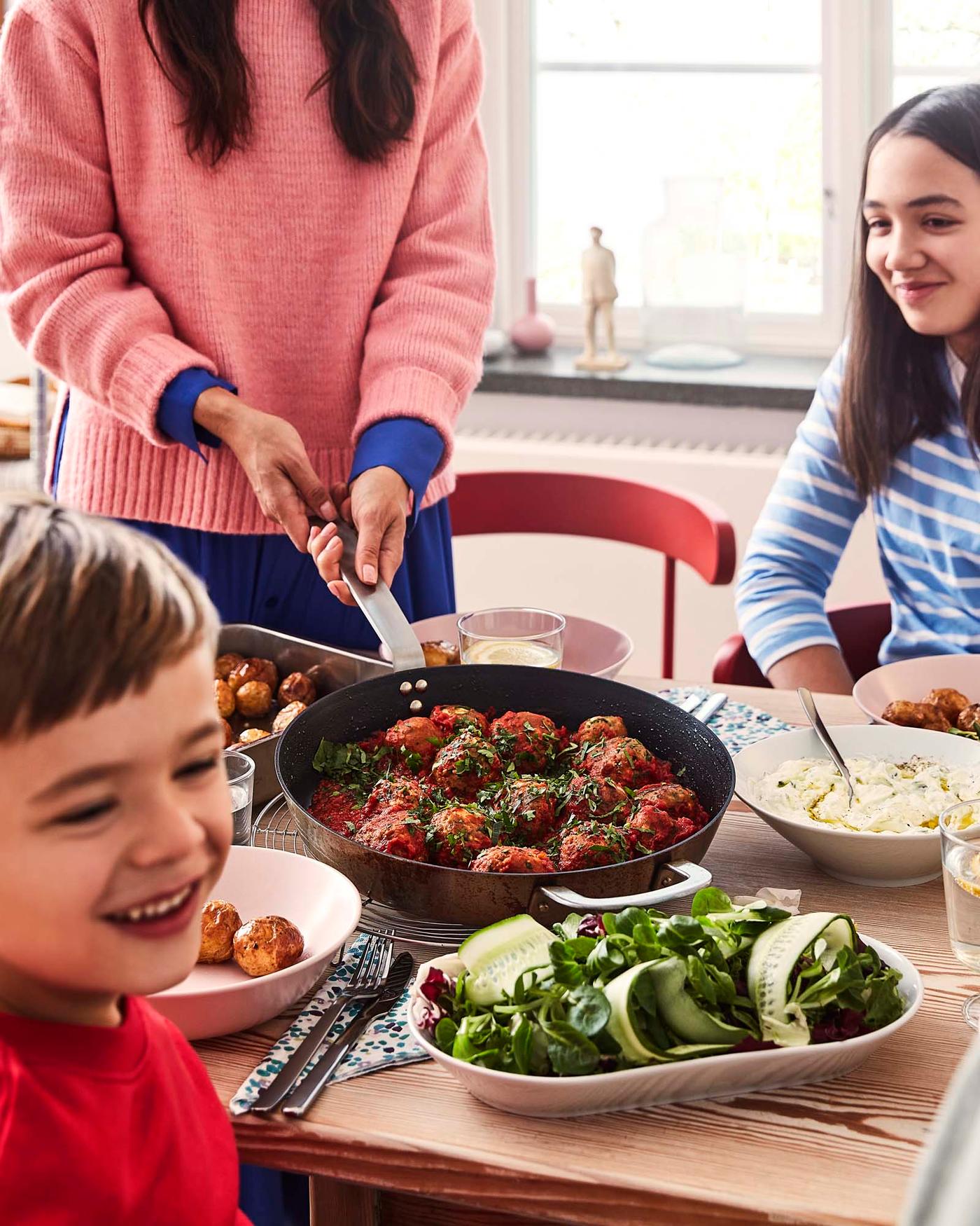 A family of three gathered at a table with a pan of plant-based meatballs and salad.