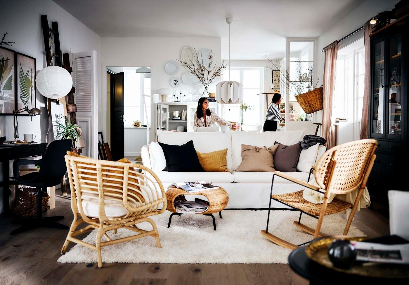 Two people in a cosy open-plan living room with a white sofa, rattan furniture, a dark wood floor, and the kitchen visible in the background.