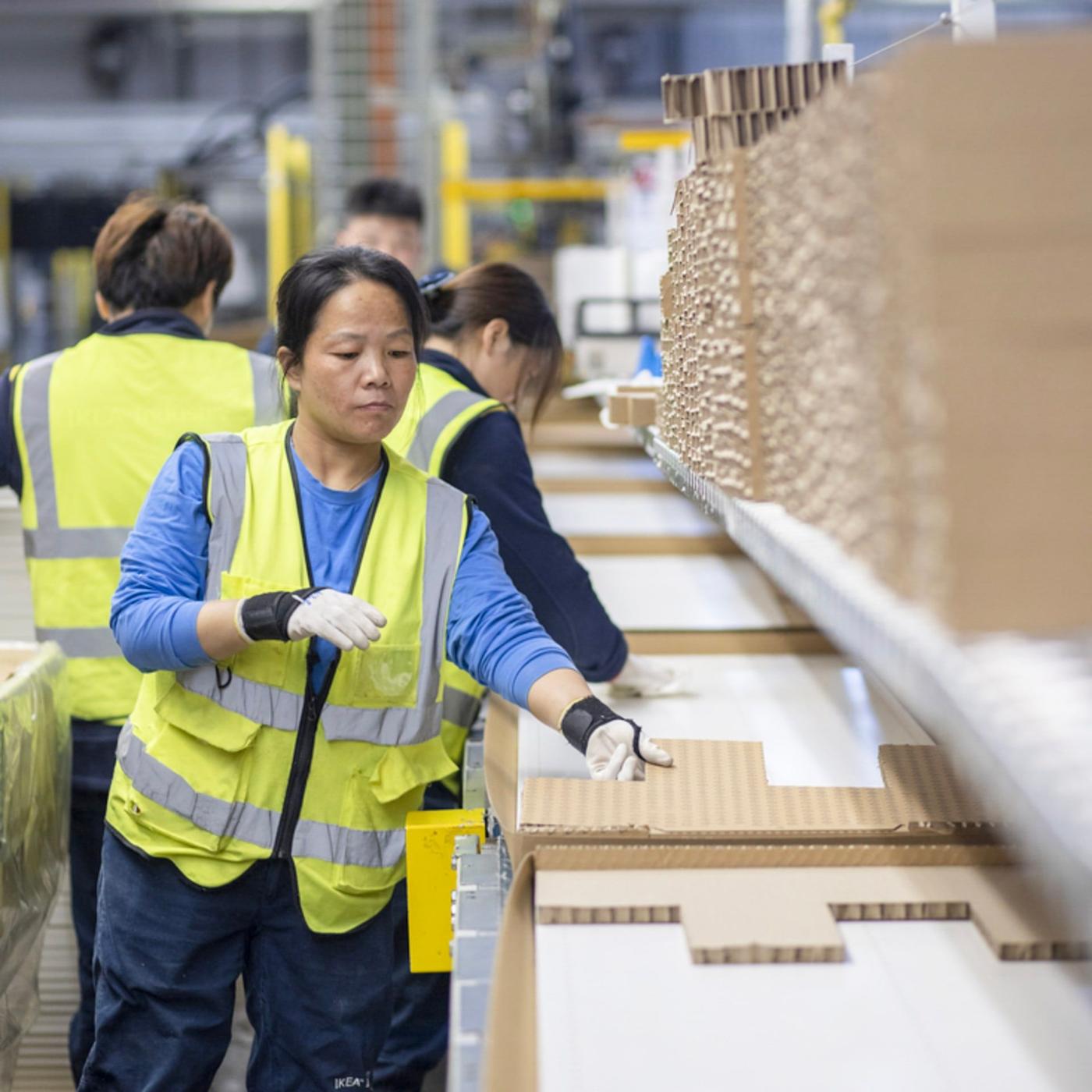 A woman in a yellow jacket standing next to a pile of cardboard packaging in an industrial environment.