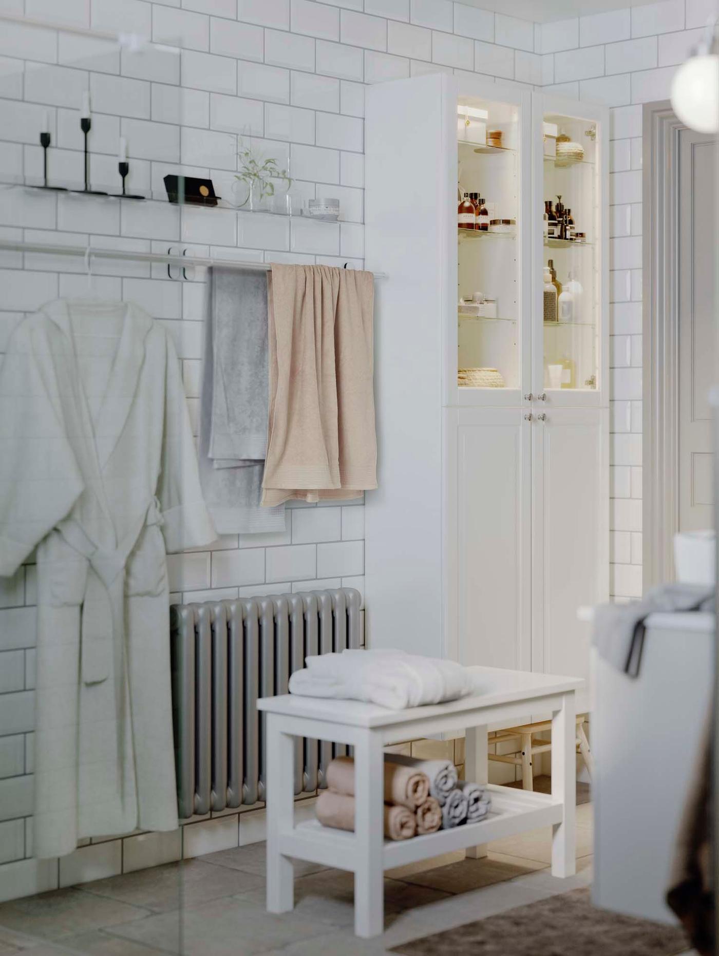 A white tiled bathroom. A robe hangs from a rail and a white bench stands in front of a radiator.