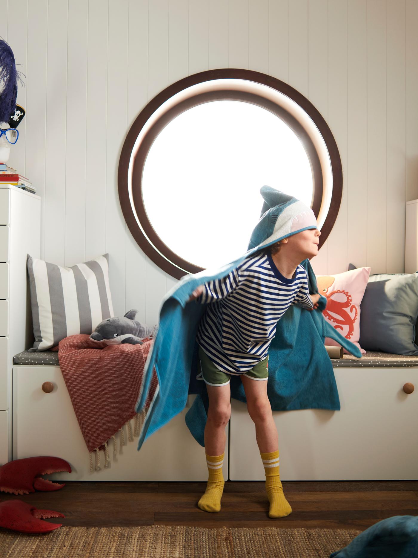 A boy standing in a room with a round shaped window and a white wooden couch in the back. On his head he's wearing the BLÅVINGAD towel, 'pretending' that he's a shark.