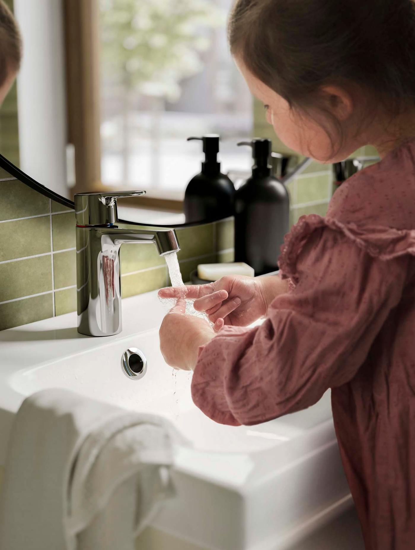 A child at a basin washing their hands under a running  chrome-plated BROGRUND wash-basin mixer tap with strainer.