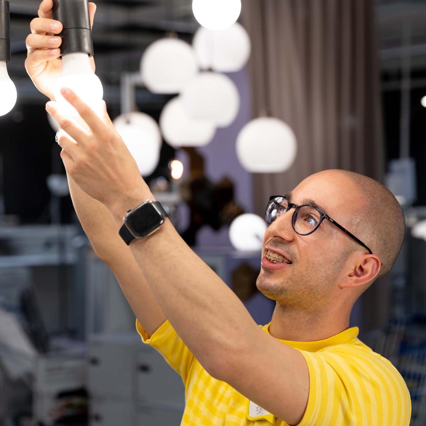 An IKEA co-worker screws an illuminated light bulb into a light bulb socket, with lights in the background.