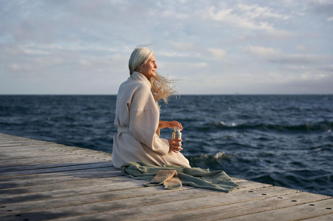 A person wearing a beige BJÄLVEN bath robe sitting on a wooden pier, looking out across a body of deep blue water.