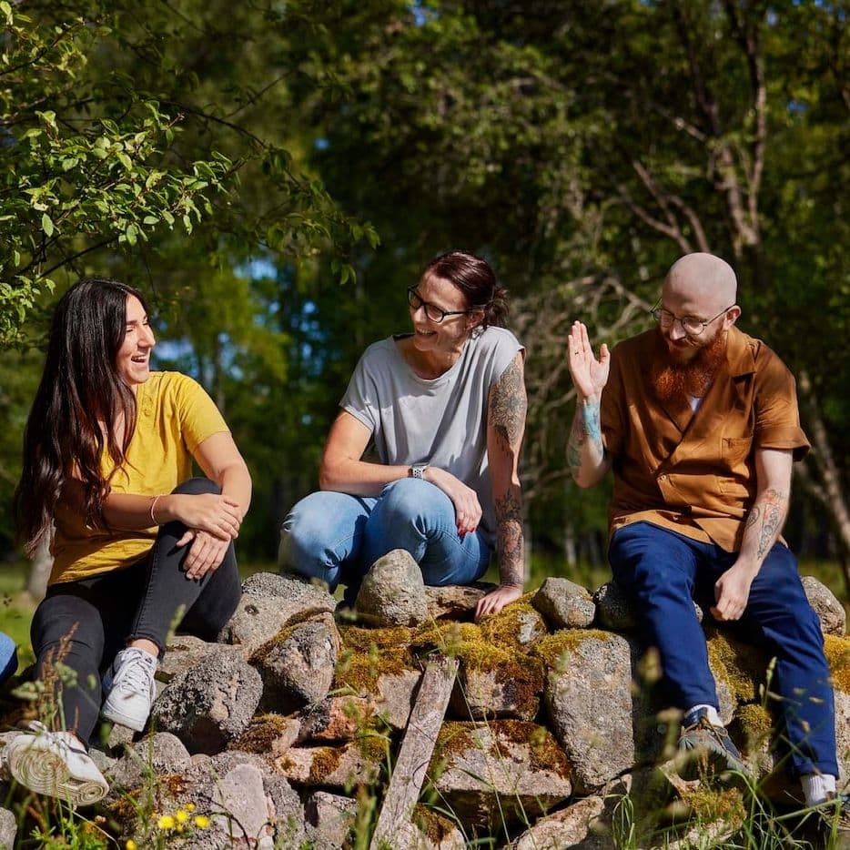 Three people sit on a stone wall near a forest. They are talking and laughing.