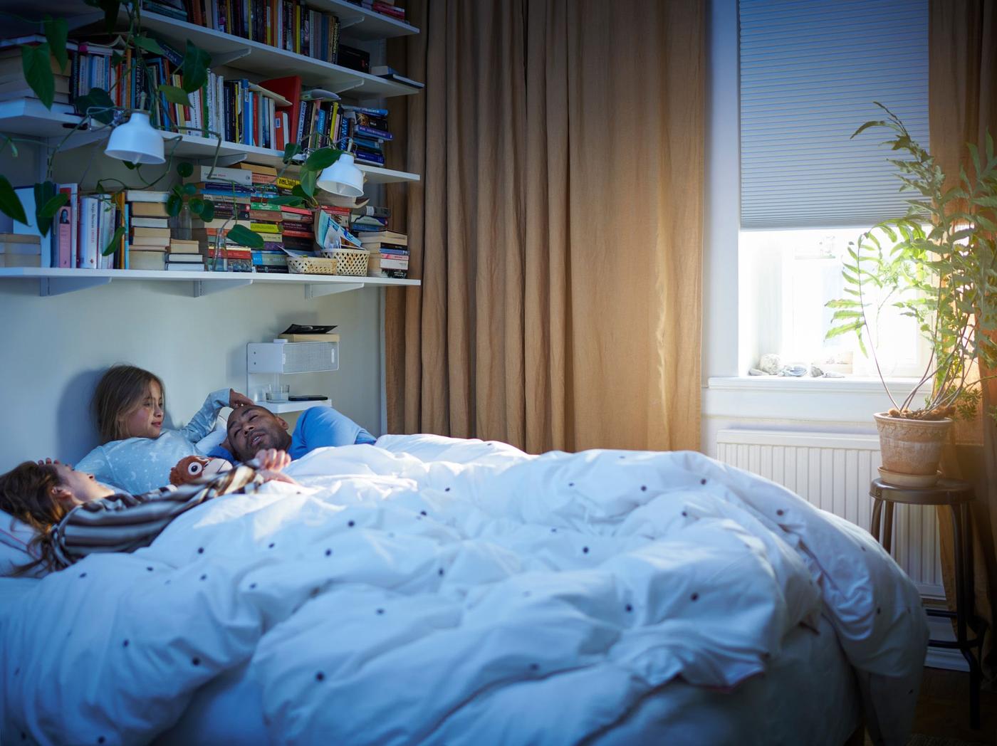 A bedroom with a child sat between two adults lying in a bed dressed in a dot pattern white/pink VÄNKRETS duvet cover and pillowcase.