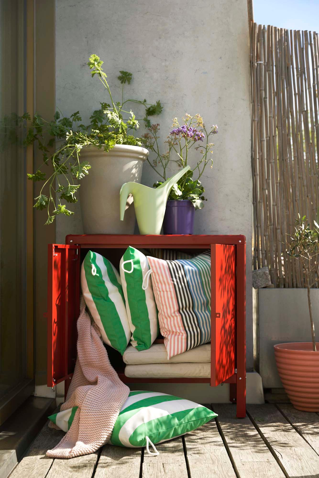 An outdoor area with plants on top of a brown-red in/outdoor KOLBJÖRN cabinet with cushions spilling from its open doors.