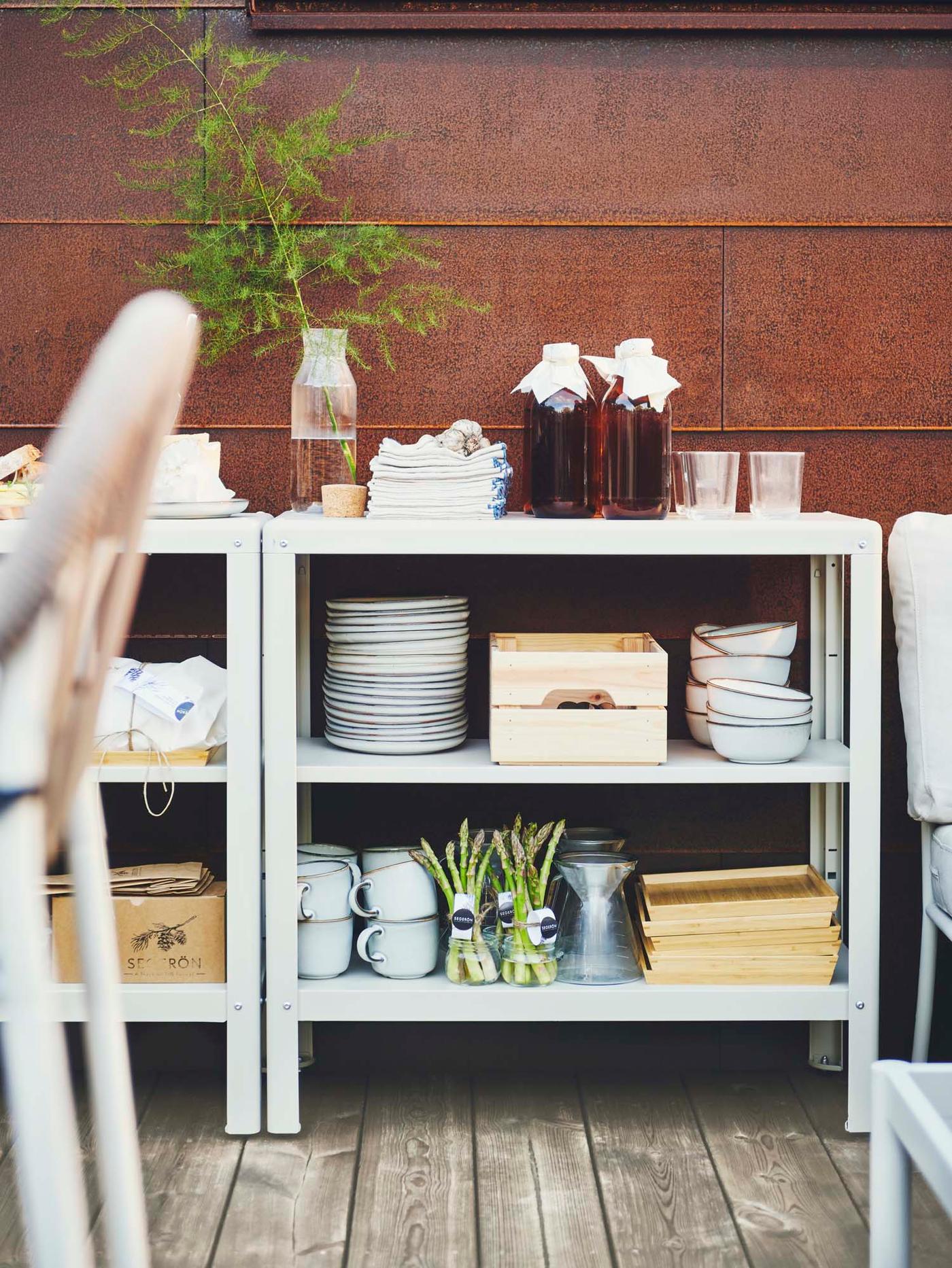 GLADELIG mugs, bowls, plates and other items on the shelves of two beige in/outdoor KOLBJÖRN shelving units.
