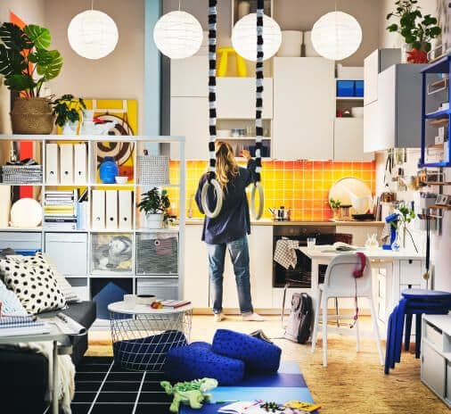 A person stands in the kitchen and living area of a colourful and compact yet generously furnished home.
