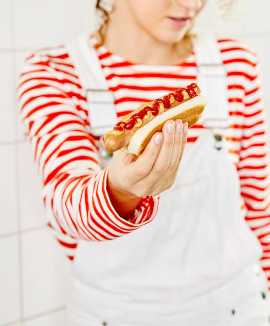 A person wearing a red and white striped shirt and white dungarees holding an IKEA plant-based hotdog.