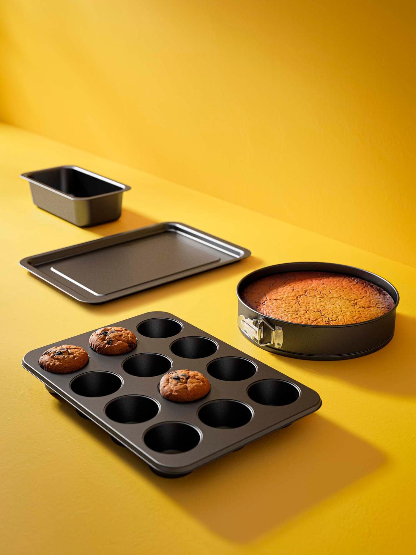 A loaf tin, baking tray, muffin pan and cake form arranged neatly on a yellow background, with baked goods in the muffin pan and cake form.