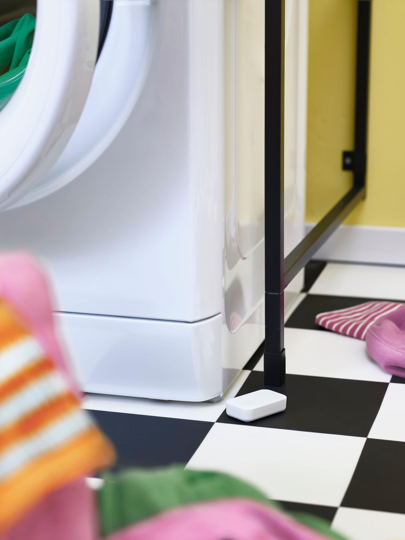 Half of a washer is standing on black and white checked floor with it's door open and a water leakage sensor lying next to it.