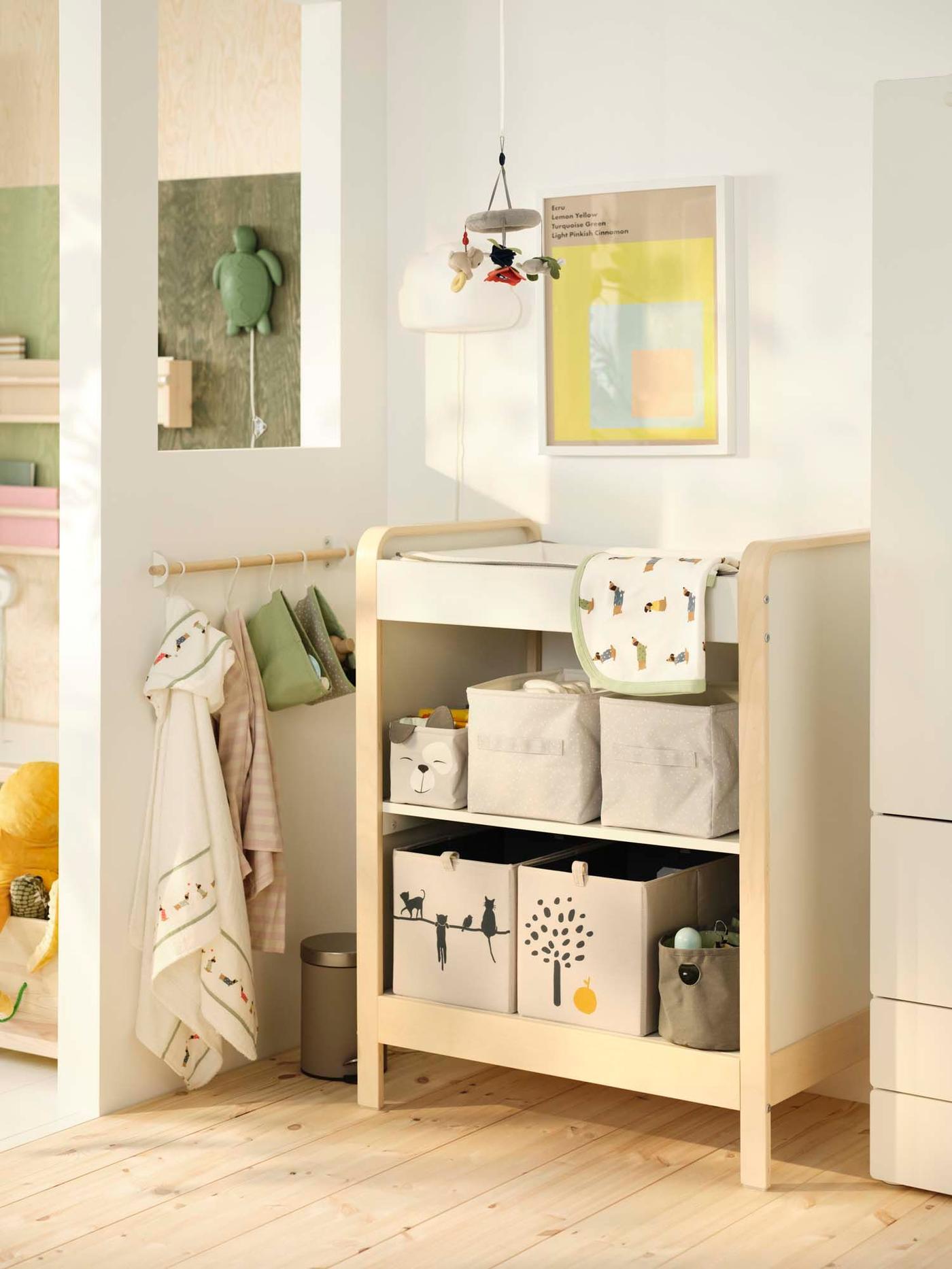 An open changing table filled with soft boxes of nappy changing supplies with a mobile above and a hanging rail next to it, in a nursery.