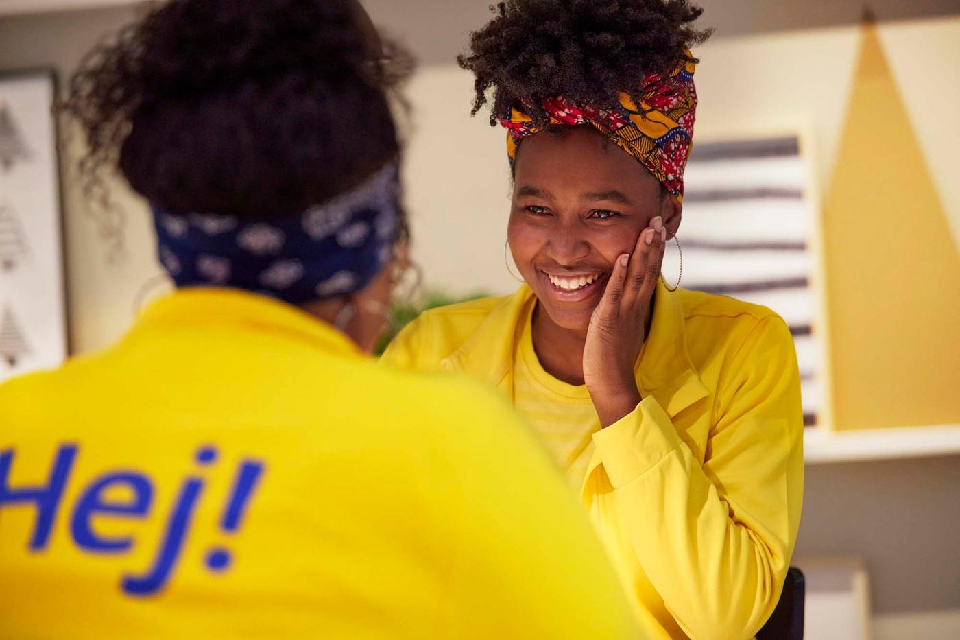 Two IKEA co-workers sit together at a table, talking and laughing. One of them has their back to the camera.